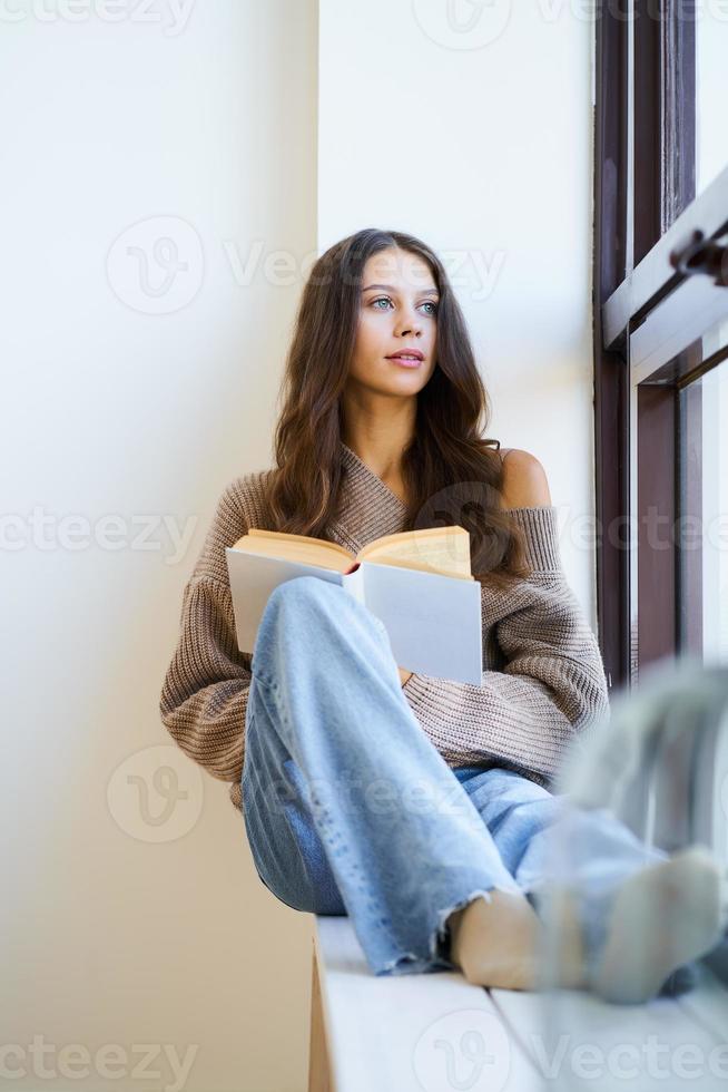 mooie jonge vrouw die een boek leest en uit het raam kijkt, alleen denkend. zelfonderdompeling foto