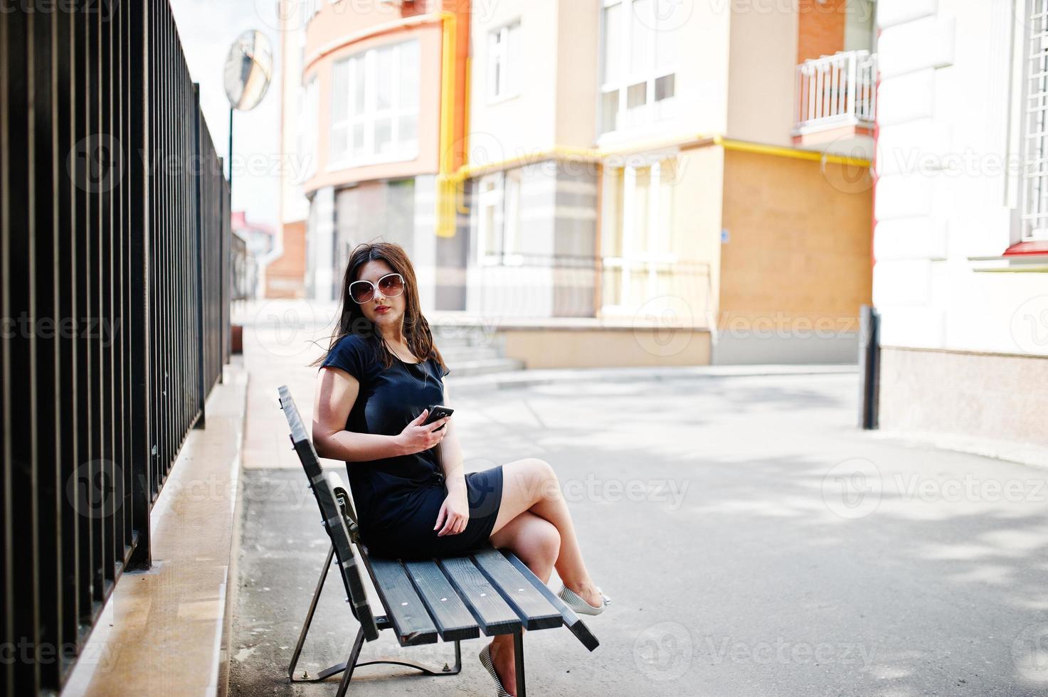 brunette meisje op zwarte jurk, zonnebril zittend op de bank, muziek luisteren via koptelefoon telefoon, en poseren op straat van de stad. foto