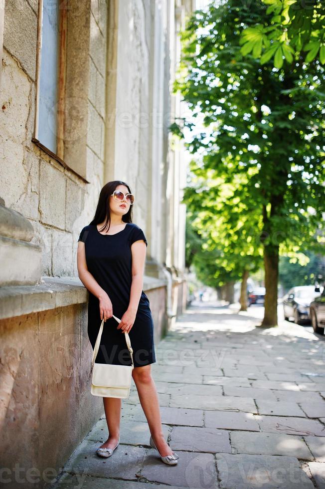 brunette meisje op zwarte jurk op zonnebril met handtas bij de hand poseren op straat van de stad. foto