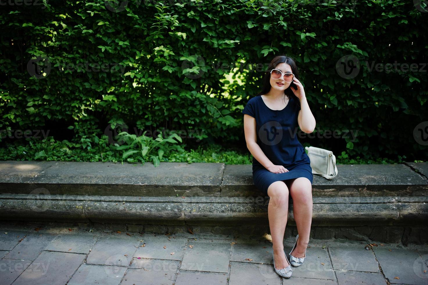 brunette zaken meisje op zwarte jurk op zonnebril zittend aan de grens tegen struiken, poseren op straat van de stad. foto