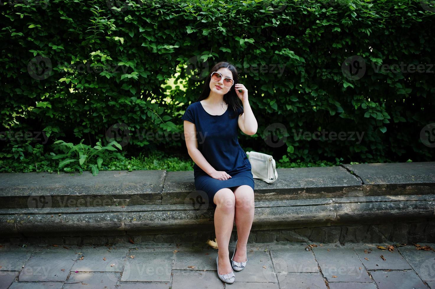 brunette zaken meisje op zwarte jurk op zonnebril zittend aan de grens tegen struiken, poseren op straat van de stad. foto