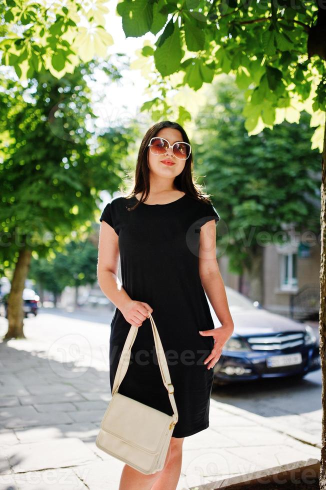 brunette business meisje op zwarte jurk op zonnebril met handtas bij de hand poseren op straat van de stad, achtergrond auto op weg. foto