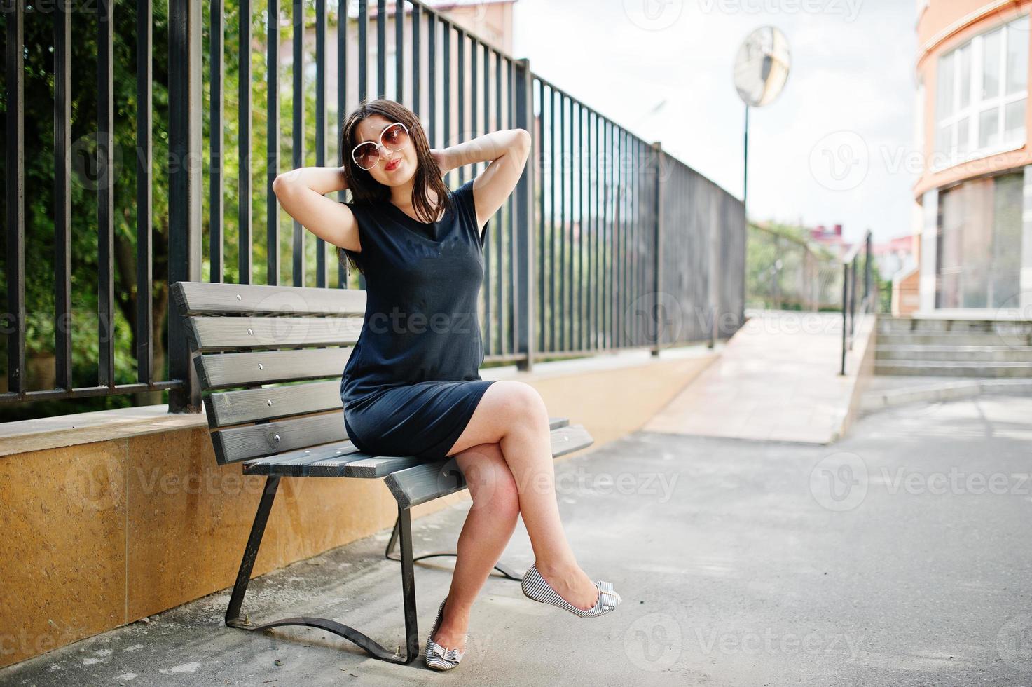 brunette meisje op zwarte jurk op zonnebril zittend op een bankje en poseren op straat van de stad. foto