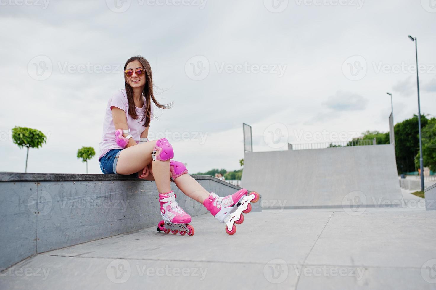 dutje code Slim portret van een aantrekkelijke jonge vrouw in korte broek, t-shirt,  zonnebril en skeelers zittend op de betonnen bank in de  buitenrolschaatsbaan. 7304914 Stockfoto