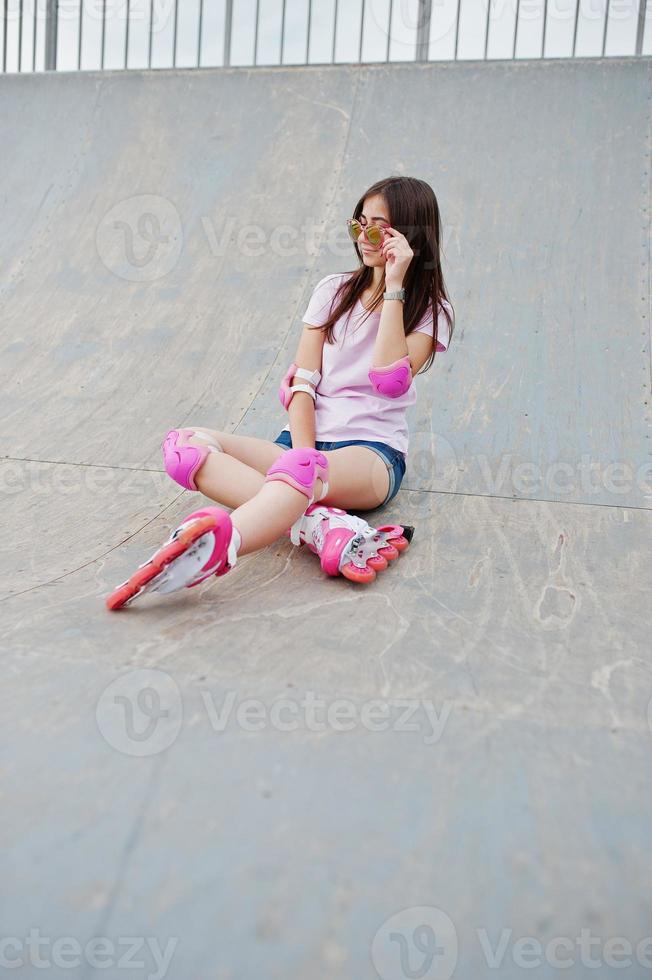 portret van een mooi meisje zittend op de skatebaan buiten in korte broek, t-shirt, zonnebril en rolschaatsen. foto