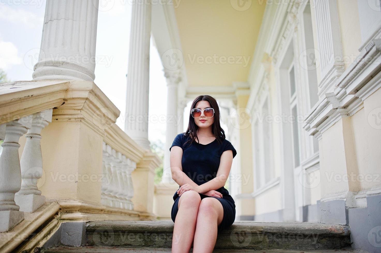 brunette meisje op zwarte jurk, zonnebril zittend op trappen van oude vintage huis, poseren op straat van de stad. foto