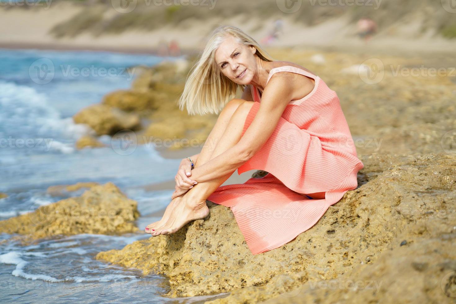 oudere vrouw zittend op een paar rotsen aan de oever van een tropisch strand, gekleed in een mooie oranje jurk. foto