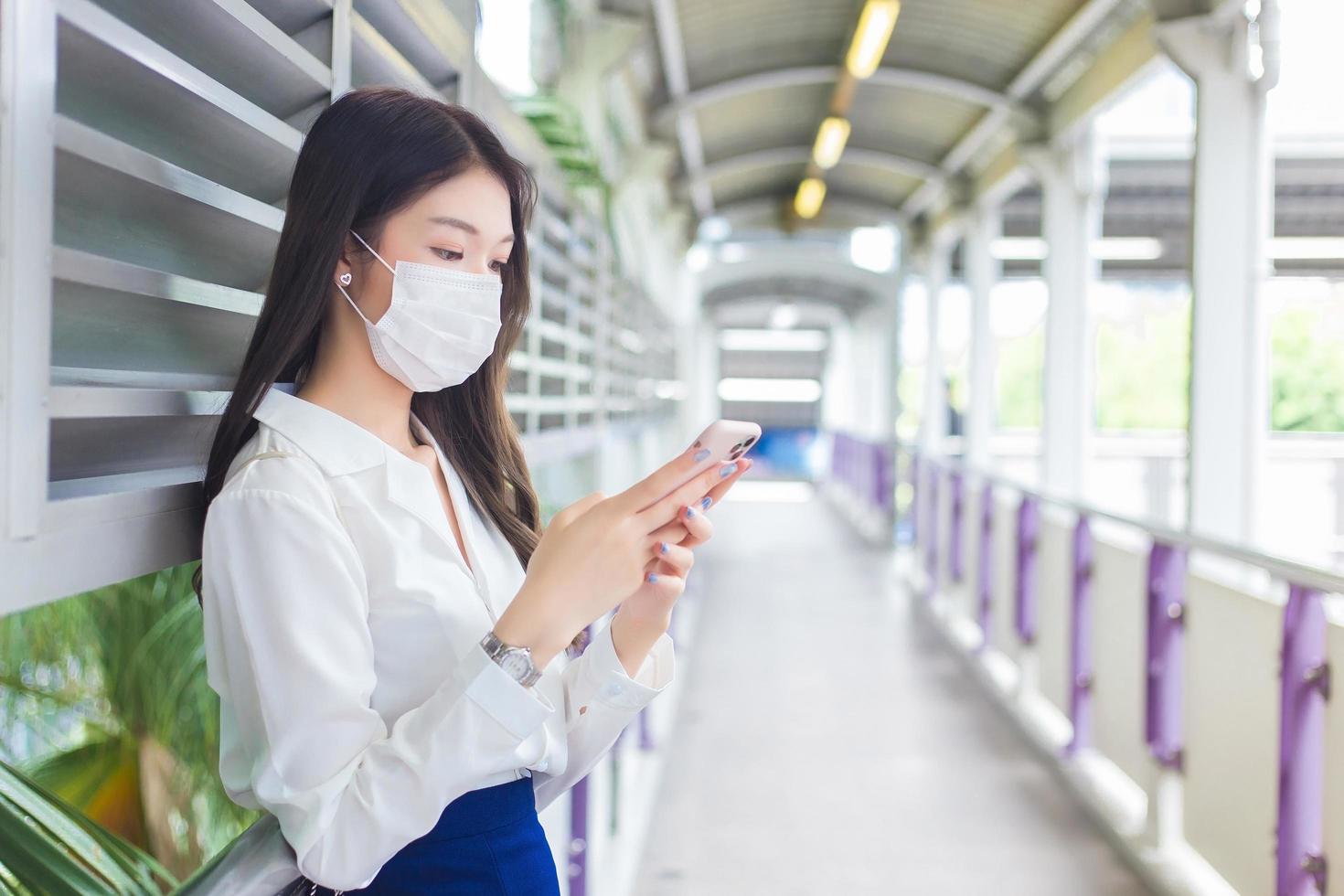 jonge aziatische mooie zakenvrouw met een gezichtsmasker staat op viaduct van skytrain in de stad terwijl ze haar smartphone gebruikt om berichten naar kantoorpersoneel te sturen. foto