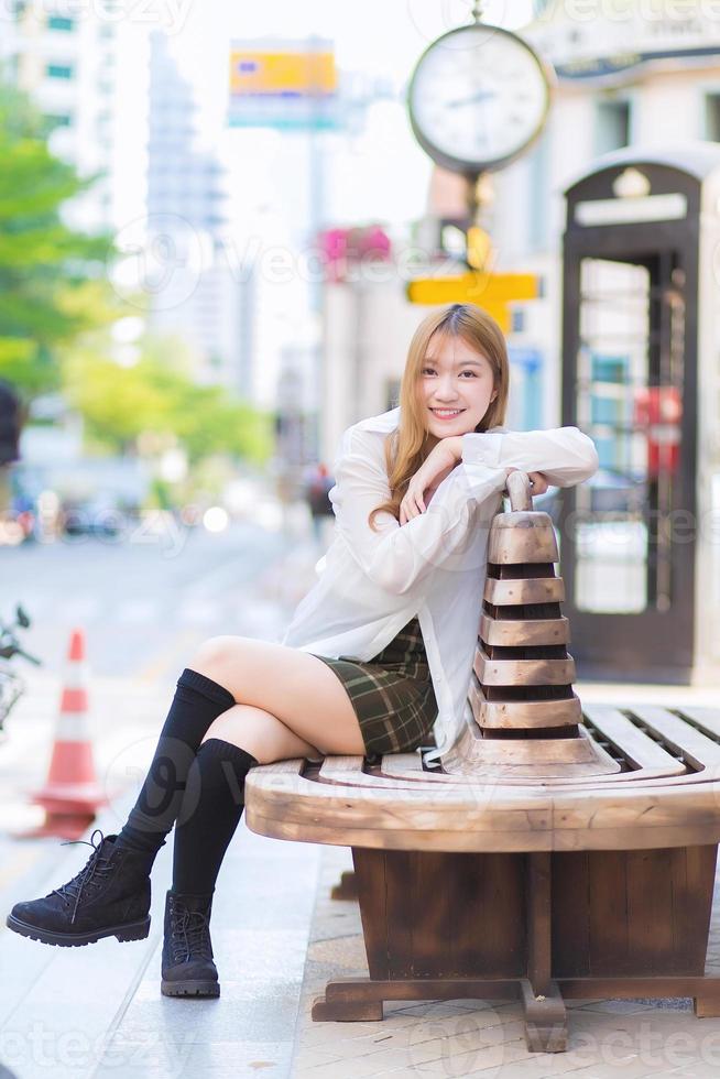 jonge aziatische mooie vrouw met bronzen haar in wit overhemd terwijl ze een gelukkige smilie aan de rand van een stadsstraat zit. foto