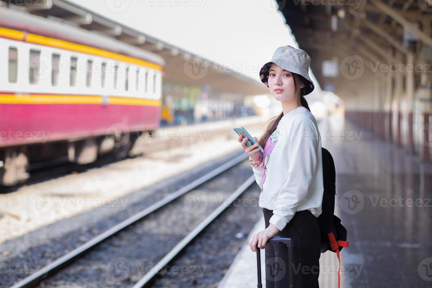 een aziatische vrouw draagt een tas en een smartphone op zoek naar reisinformatie. tijdens het wachten op de trein op het perron foto