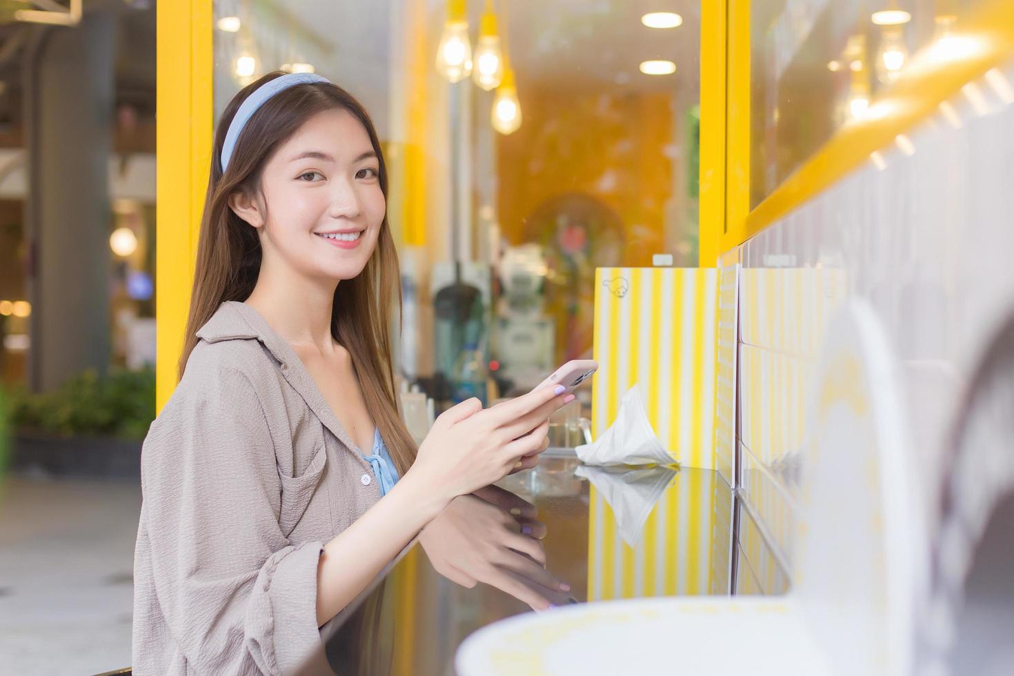 mooie jonge aziatische vrouw met lang haar kijkt naar de camera terwijl ze een smartphone vasthoudt en speelt om contact te maken met iemand in het dessertcafé in het winkelcentrum. foto