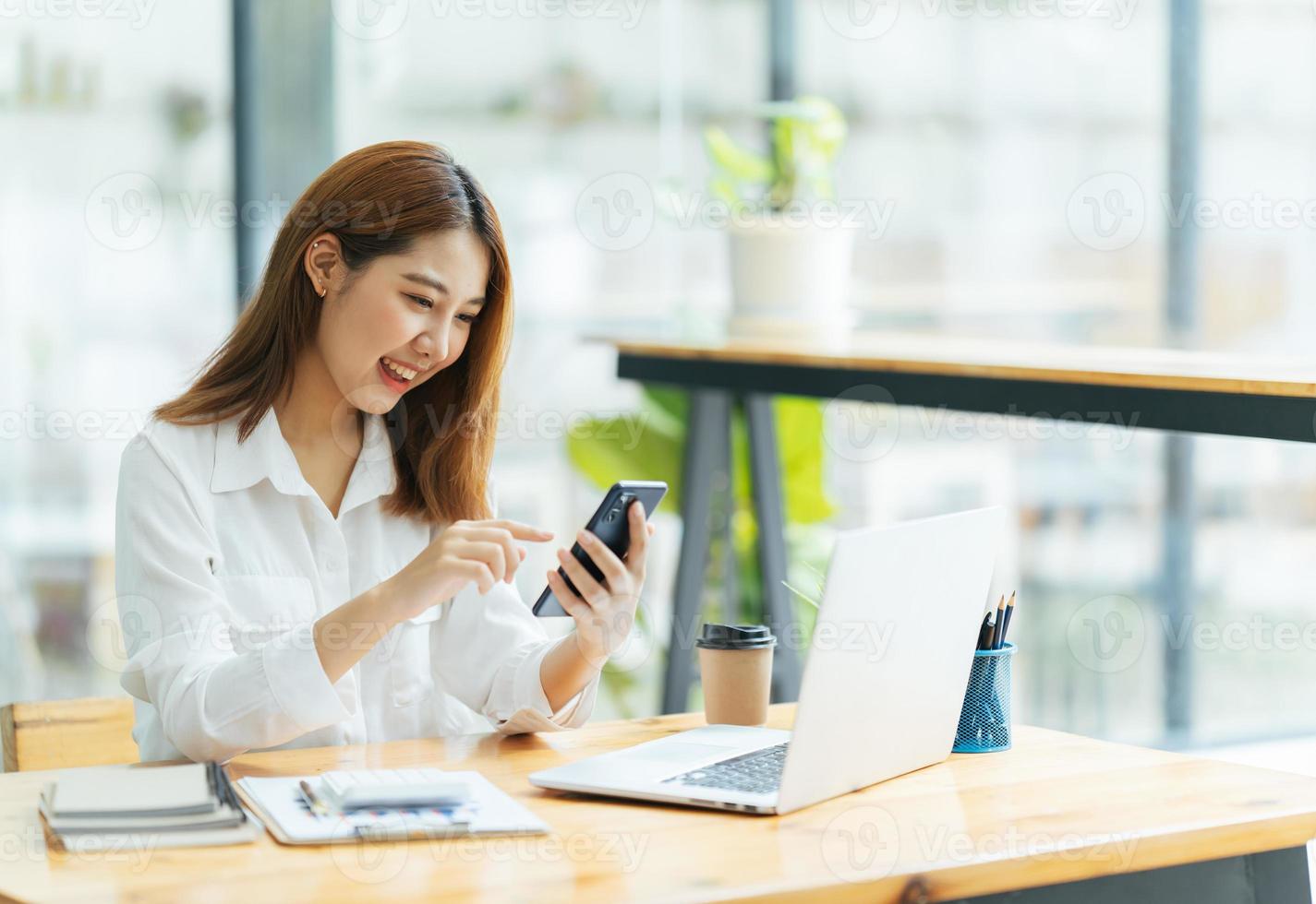 aziatische vrouw in vrijetijdskleding is blij en vrolijk terwijl ze communiceert met haar smartphone en in een coffeeshop werkt. foto