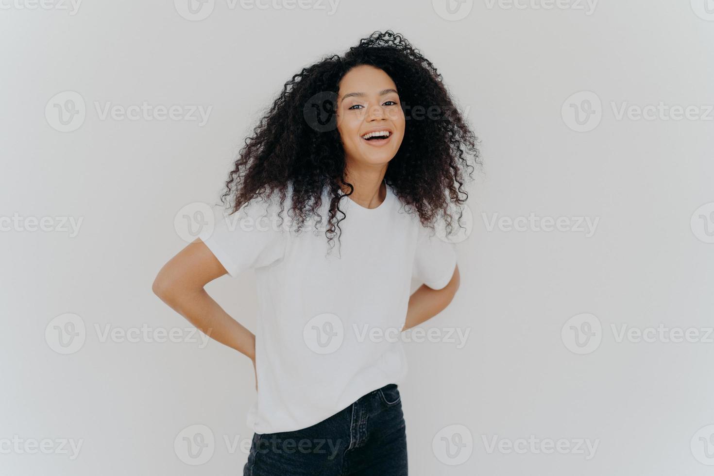 portret van vrolijke duizendjarige vrouw houdt handen achter de rug, gekleed in vrijetijdskleding, lacht om iets leuks, geïsoleerd op witte studioachtergrond. Afro-Amerikaanse dame drukt geluk uit foto