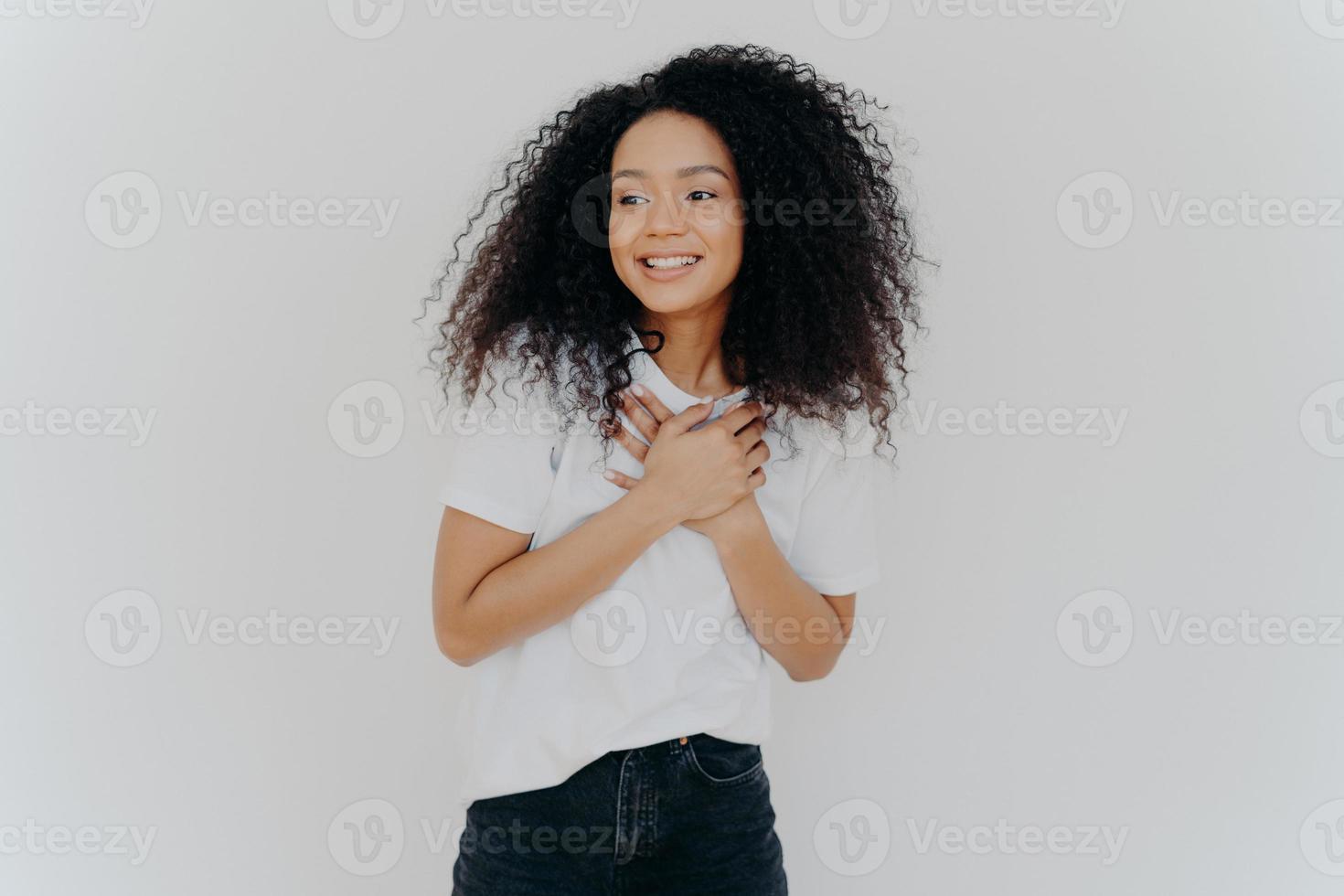 foto van gelukkig meisje houdt beide handen op de borst, kijkt opzij met een aangename glimlach, merkt iets geweldigs op, poseert tegen een witte achtergrond, draagt een casual t-shirt en een spijkerbroek. positieve emoties concept