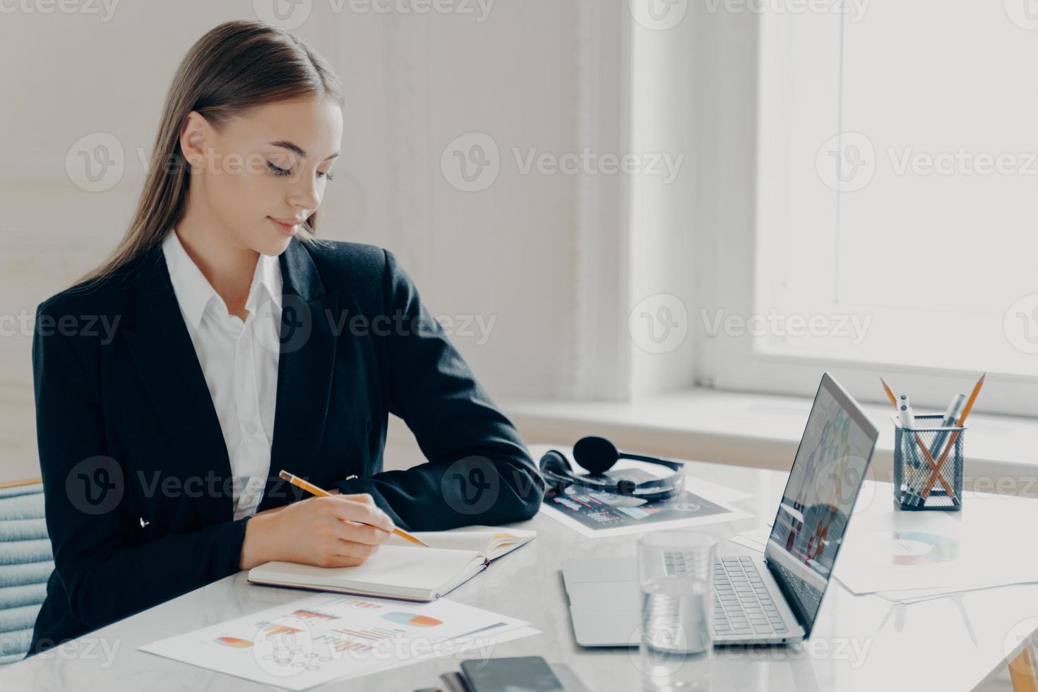 zakenvrouw die bij het bureau zit en in een notitieboek voor de laptop schrijft foto