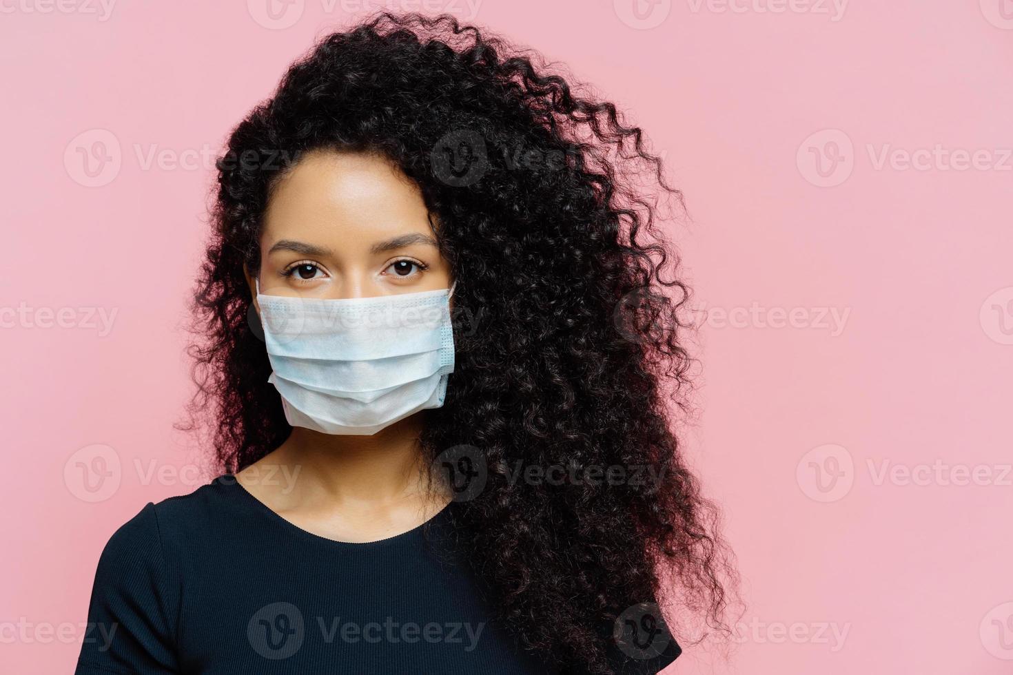 ernstige jonge vrouw met een donkere huid die thuis in zelfisolatie zit, een beschermend medisch masker draagt, thuis in quarantaine is, nonchalant gekleed is, poseert tegen een roze achtergrond. coronavirusziekte foto