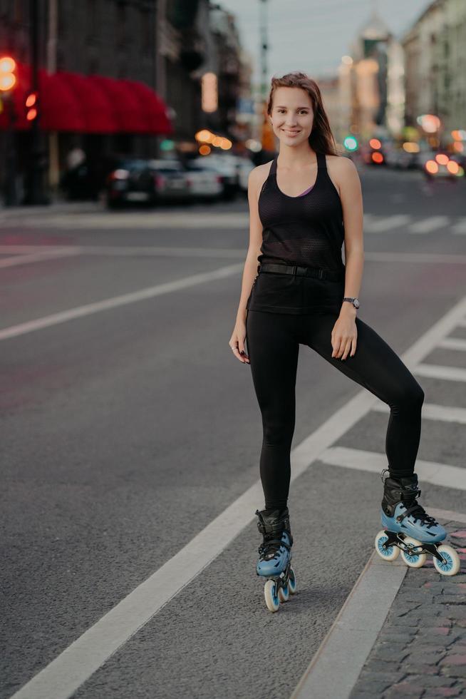 volledige lengte shot van actieve jonge vrouw in goede fysieke conditie gekleed in zwarte activewear geniet van skaten tijdens goede zomerdag poses op weg tegen de achtergrond van de stad. recreatie. foto