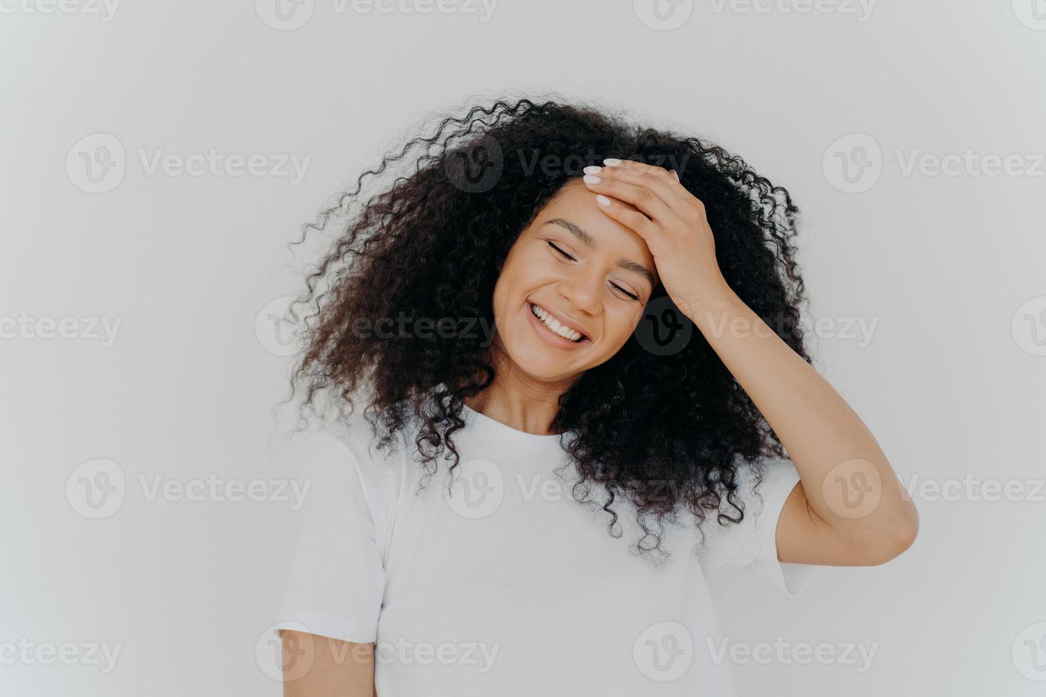 foto van gelukkige dame houdt hand op voorhoofd, houdt hoofd schuin, glimlacht vrolijk, houdt ogen gesloten, heeft afro-krullend haar, draagt casual wit t-shirt, geniet van een aangenaam moment in het leven, voelt zich een beetje verlegen