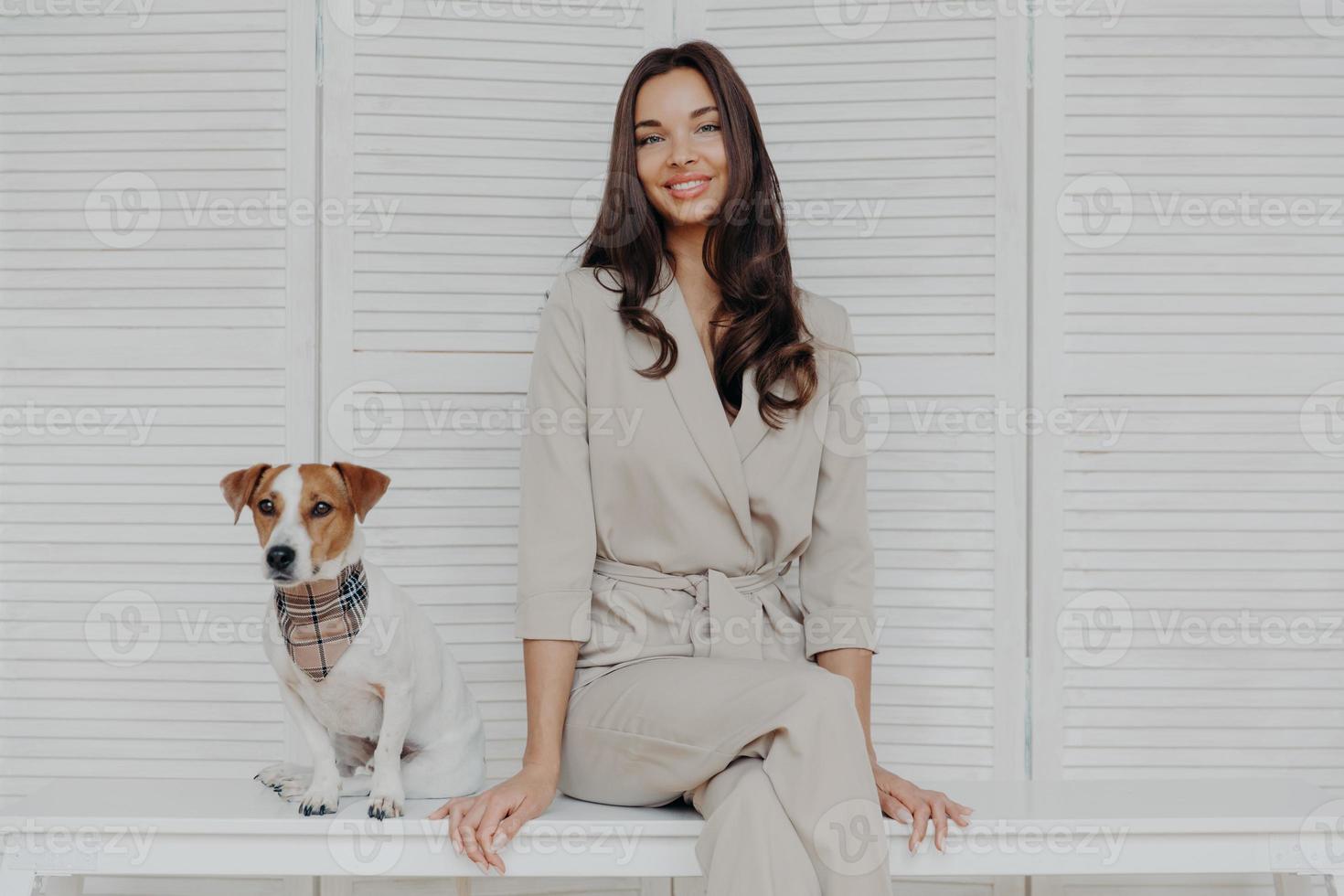 afbeelding van aantrekkelijke brunette Europese vrouw zit samen met rashond, gekleed in modieuze kleding, kijkt met zachte glimlach op camera. mensen, dierenzorg en verantwoordelijkheidsconcept foto