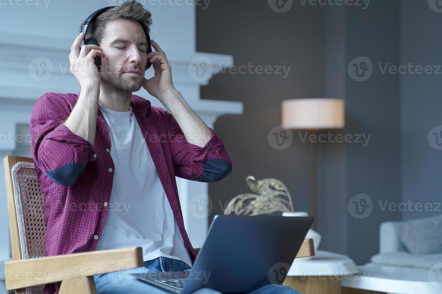 jonge duitse man zit op een rieten stoel met een laptop op schoot en luistert naar muziek in een koptelefoon foto