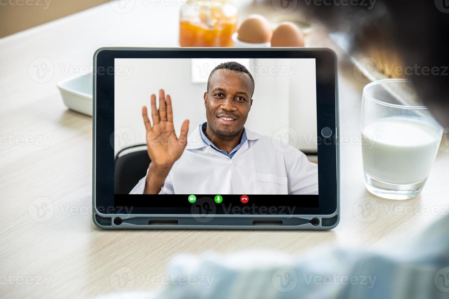 close-up van een Afro-Amerikaanse vader die zwaait terwijl hij via een videogesprek communiceert met zijn dochter die hem belt terwijl hij ochtendmelk van huis heeft. foto