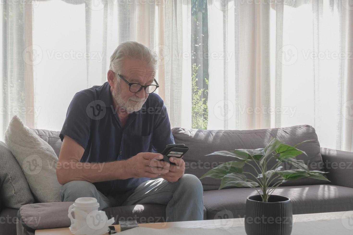 lifestyle fotografie van blanke oude man die alleen woont in vakantie, met behulp van smartphone, sigaret en koffiekopje op tafel gezet in woonkamer gezellig huis en zonnige dag. foto