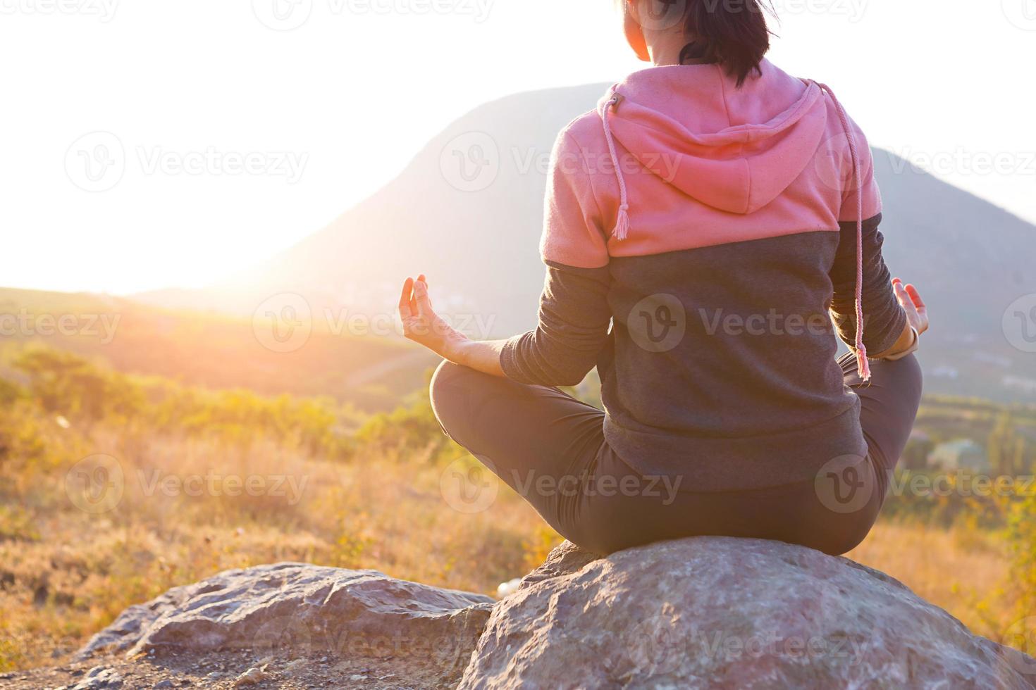 een vrouw zit op een rots in lotushouding met haar vingers gesloten in een teken van harmonie, ontspanning en meditatie. yoga, zelfonderdompeling, balans met de natuur. bergen, zonlicht. kopieer ruimte foto