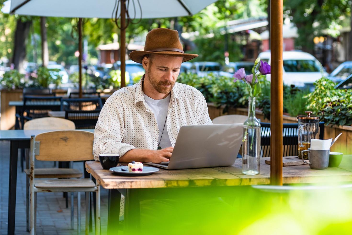jonge stijlvolle hipster in hoed werkt met laptop buiten in het straatcafé foto