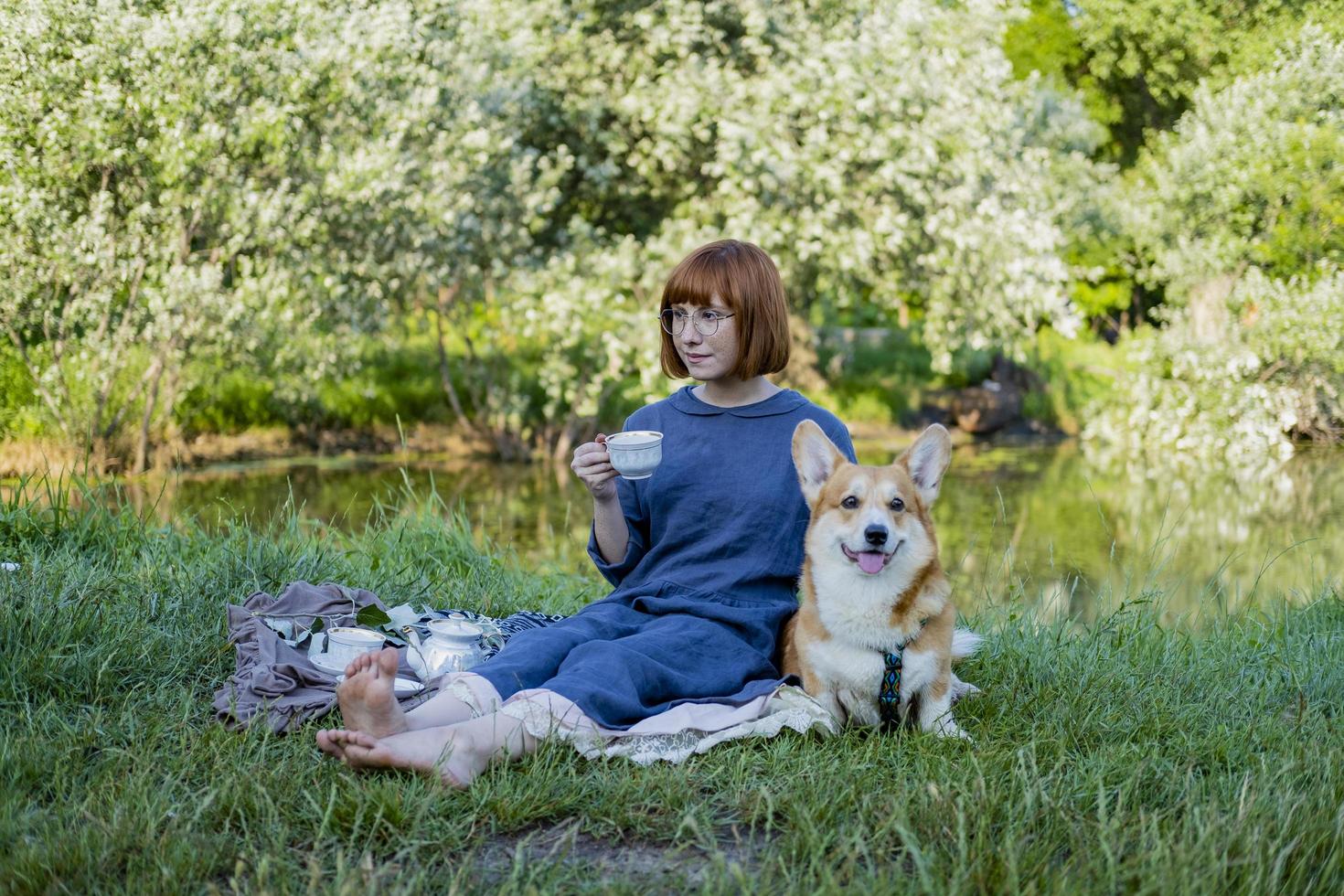 jonge vrouw in retro jurk met grappige corgi-hond op de picknick, vrouw met schattige hond drinkt Engelse thee in het park foto