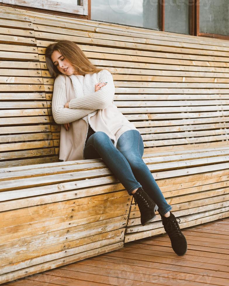 mooi jong meisje met lang bruin haar zit op houten bank gemaakt van planken en rust, slaapt of dommelt in de frisse lucht. outdoor fotoshoot met aantrekkelijke vrouw in de winter of herfst. foto