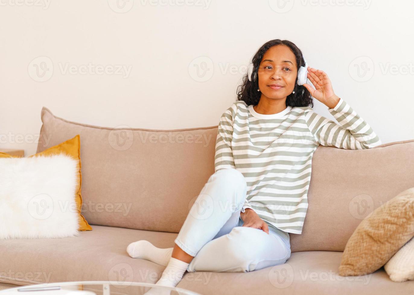 lachende Afro-Amerikaanse vrouw die naar muziek luistert in een koptelefoon terwijl ze op de bank zit foto