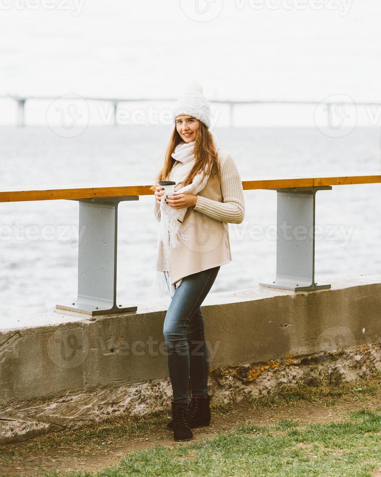 mooi jong meisje koffie, thee drinken uit plastic mok in de herfst, winter. een vrouw met lang haar staat aan de waterkant aan de Oostzee in de haven en wacht op de veerboot, verwarmd door een warme drank, kopieer ruimte foto