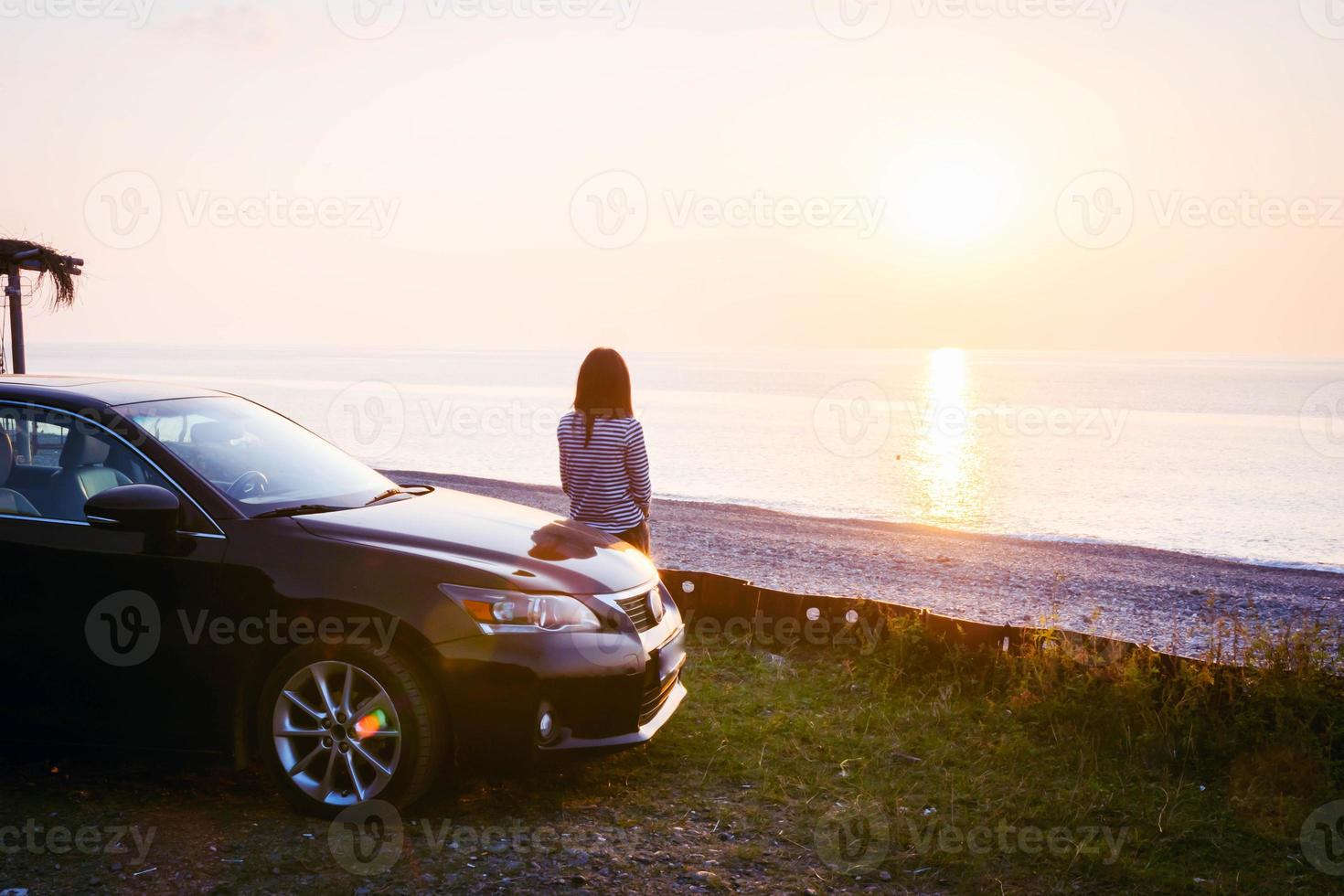 vrouw staat bij de zwarte luxe auto en kijkt naar een panoramisch uitzicht op de kalme zee en de ondergaande zon. tsikhisdziri. Georgië. roadtrip-concept en solo-ontspanning aan zee-concept. foto