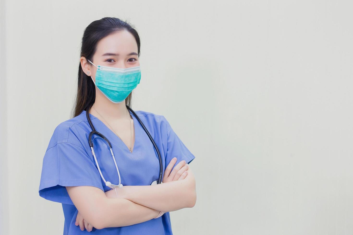 aziatische vrouw arts draagt medisch uniform met stethoscoop en medisch gezichtsmasker, arm oversteken op witte achtergrond. foto