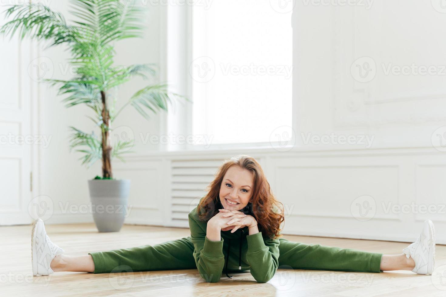 fitnessvrouw demonstreert mooie flexibiliteit, doet gymnastiekoefeningen, toont beensplitsing, houdt handen onder de kin, glimlacht breed, poseert op de vloer in een ruime kamer. mensen, training, aerobicsconcept foto