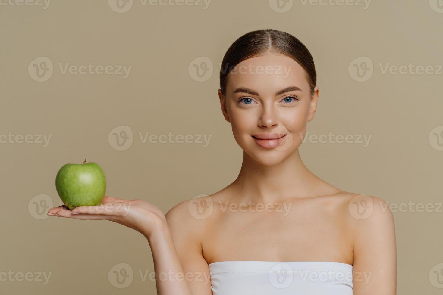 foto van gezonde, mooie vrouw met natuurlijke make-upstandaard gewikkeld in badhanddoek houdt groene appel aan beveelt product voor diëten aan geïsoleerd op bruine achtergrond maakt natuurlijke zelfgemaakte gezichtsmaskers