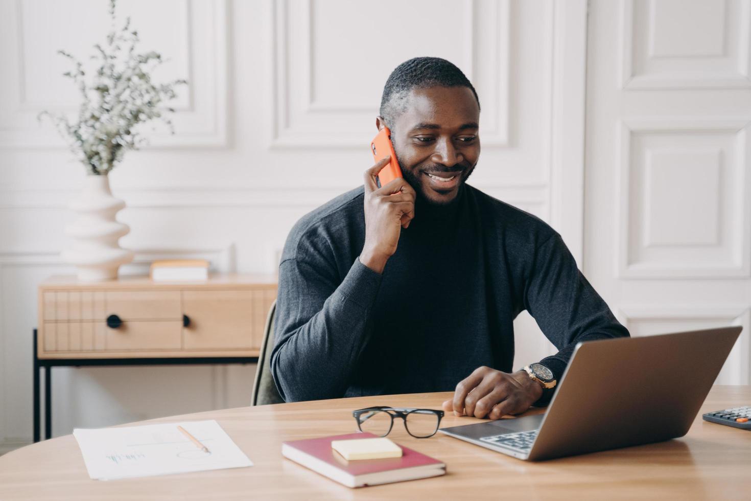 succesvolle afro-amerikaanse zakenman die een aangenaam telefoongesprek heeft terwijl hij op een laptop werkt foto