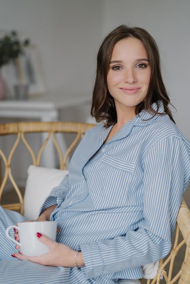 tevreden mooie vrouw met aangename uitstraling, draagt gestreepte nachtkleding, drinkt aromatische koffie, poseert binnen. ontspannen huisvrouw drinkt 's ochtends na het ontwaken. mensen en rust concept foto