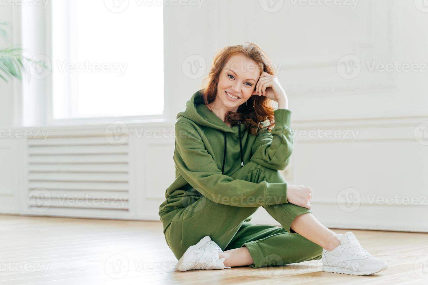 positief roodharig fitnessmeisje beoefent een gezonde levensstijl, heeft rust na binnentraining, voelt zich opgewonden na actieve oefeningen, draagt een comfortabel groen trainingspak, poseert op de vloer. fysieke activiteit foto