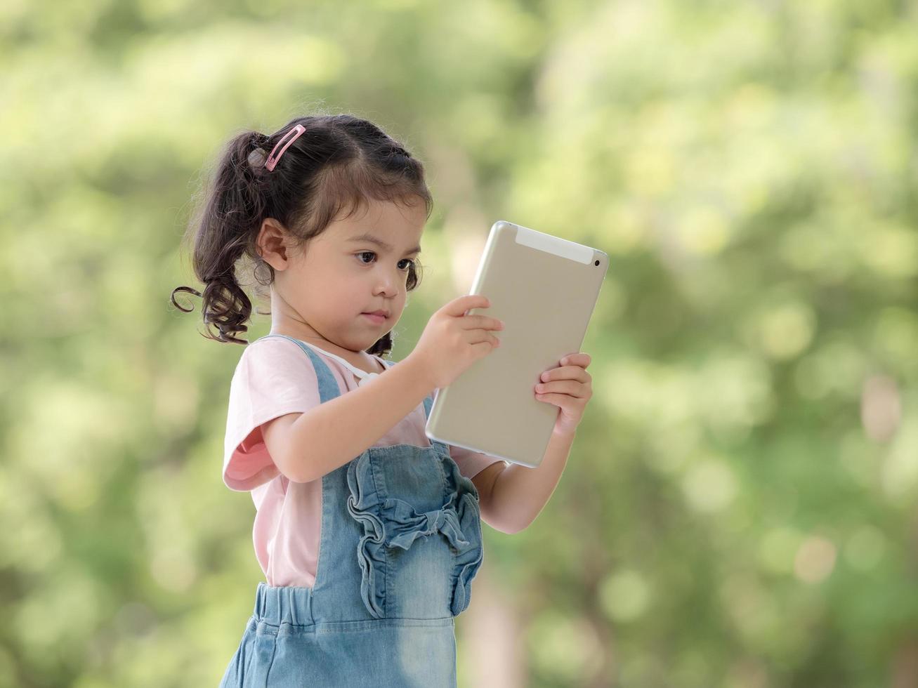 een schattig Aziatisch meisje gebruikt een tablet om spelletjes te spelen en buiten school in het park te leren foto