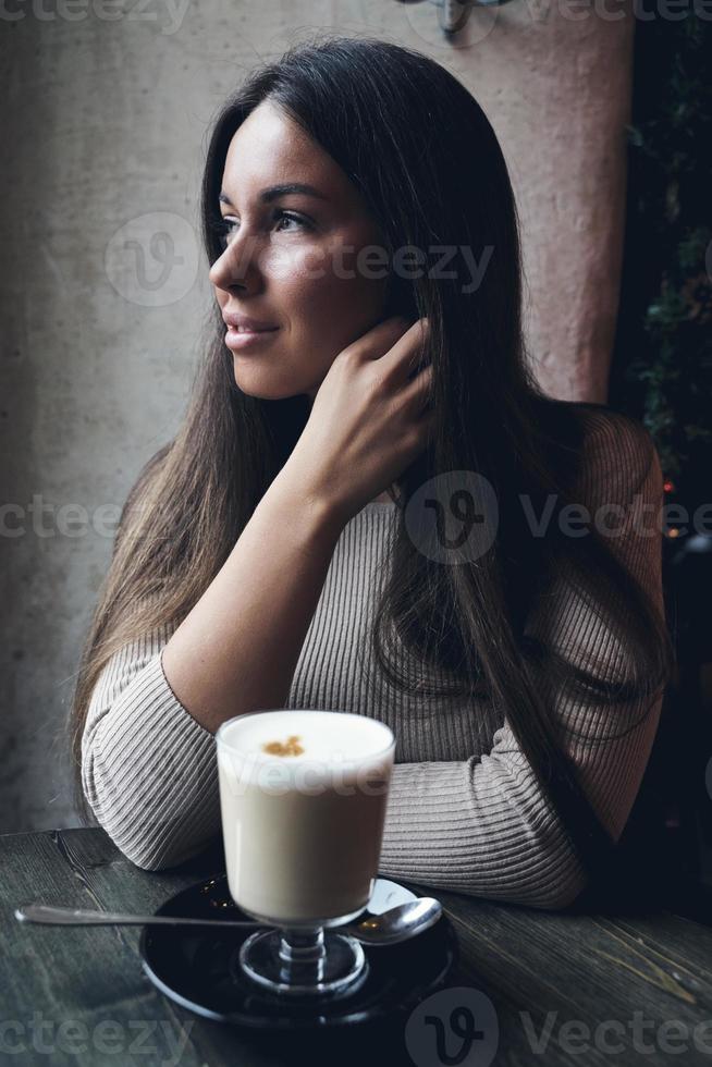 mooi meisje zit in café in kerstvakantie, in achtergrondverlichting van slinger. brunette vrouw met lang haar drinkt cappuccino koffie, latte en kijkt uit raam, donkere achtergrond foto