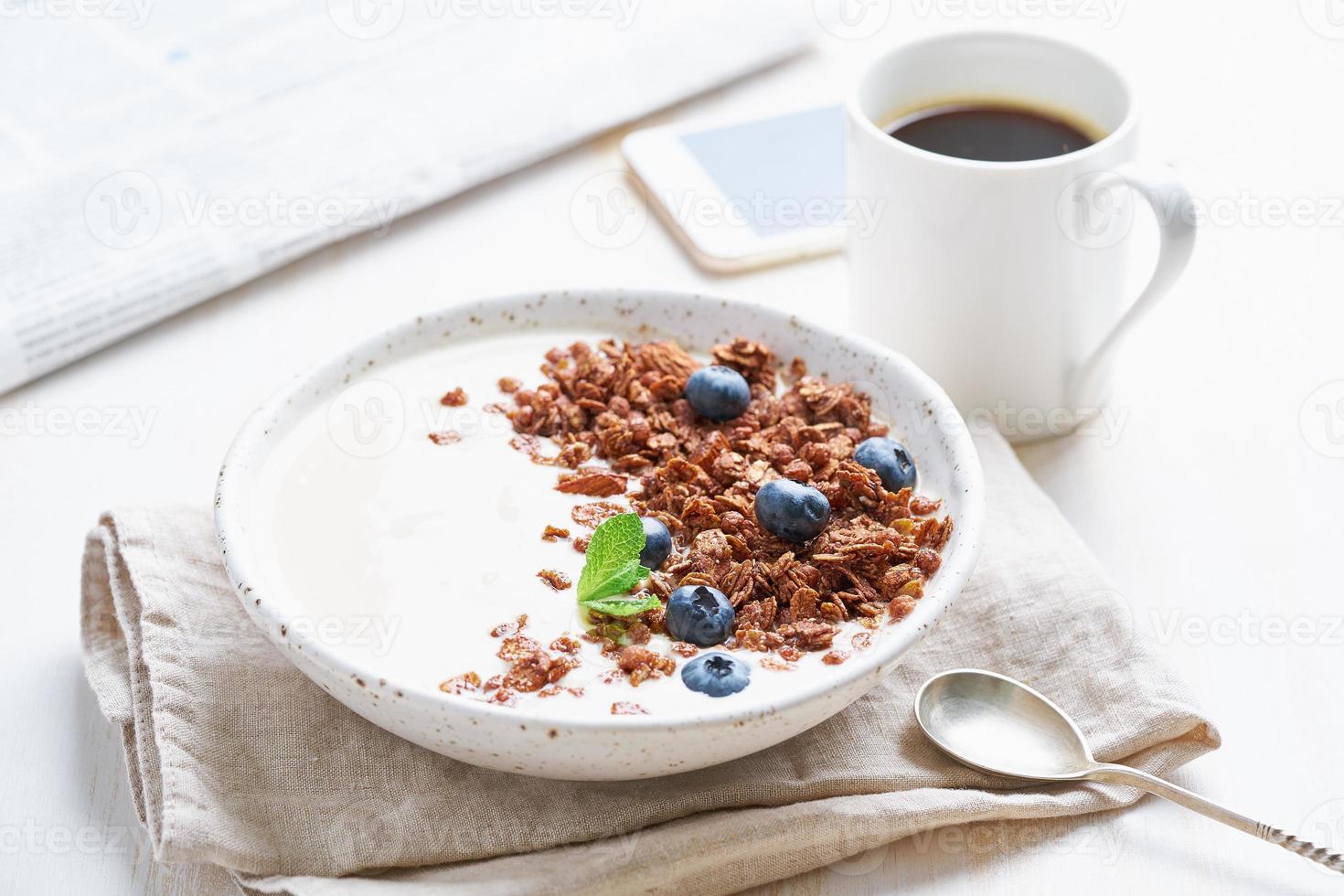 yoghurt met chocolade granola, bosbes. ontbijt met kopje koffie, mobiel, krant foto