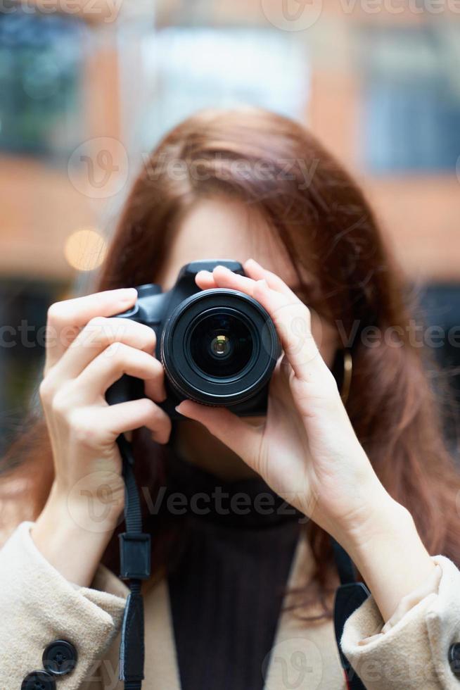 selectieve focuson lens. mooie stijlvolle modieuze meid houdt camera in haar handen en maakt foto's. vrouw fotograaf met lang donker haar in de stad, stedelijke shoot, verticaal. onherkenbaar persoon foto