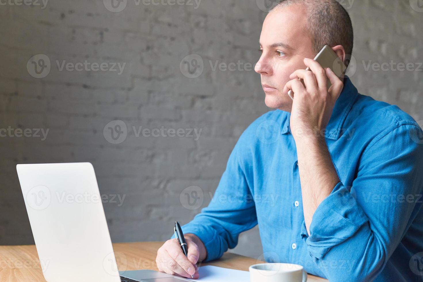 knappe knappe volwassen man kijkt uit raam en praten op mobiele telefoon, zittend op de computer, laptop. man met vrijetijdskleding in blauw shirt en tafel op kantoor voor raam, kopieer ruimte foto