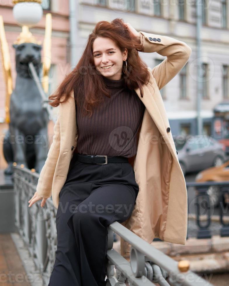 mooie serieuze stijlvolle modieuze slimme meid die op de brug staat en glimlacht, st. petersburgse stad. charmante bedachtzame vrouw met lang donker haar, topshoot. foto