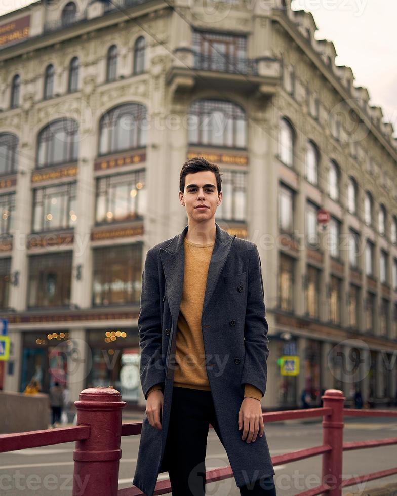 knappe stijlvolle modieuze man, brunette in elegante grijze jas, staat op straat in het historische centrum van st. petersburg. jonge man met donker haar, dikke wenkbrauwen. foto