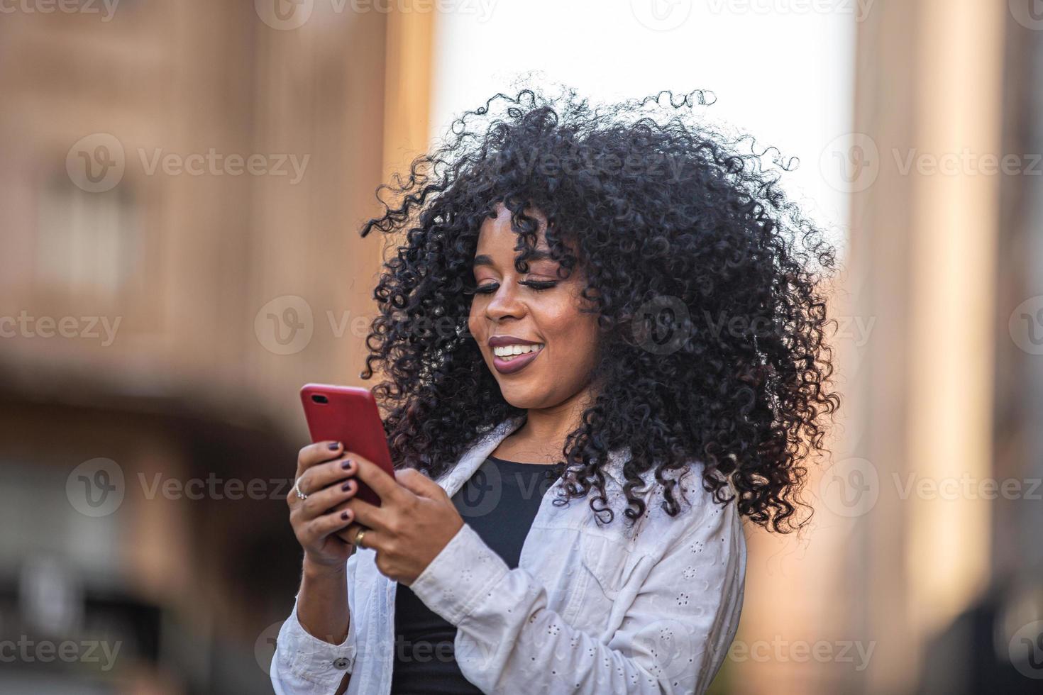 jonge krullend haar zwarte vrouw lopen met behulp van mobiele telefoon. sms'en op straat. grote stad. foto