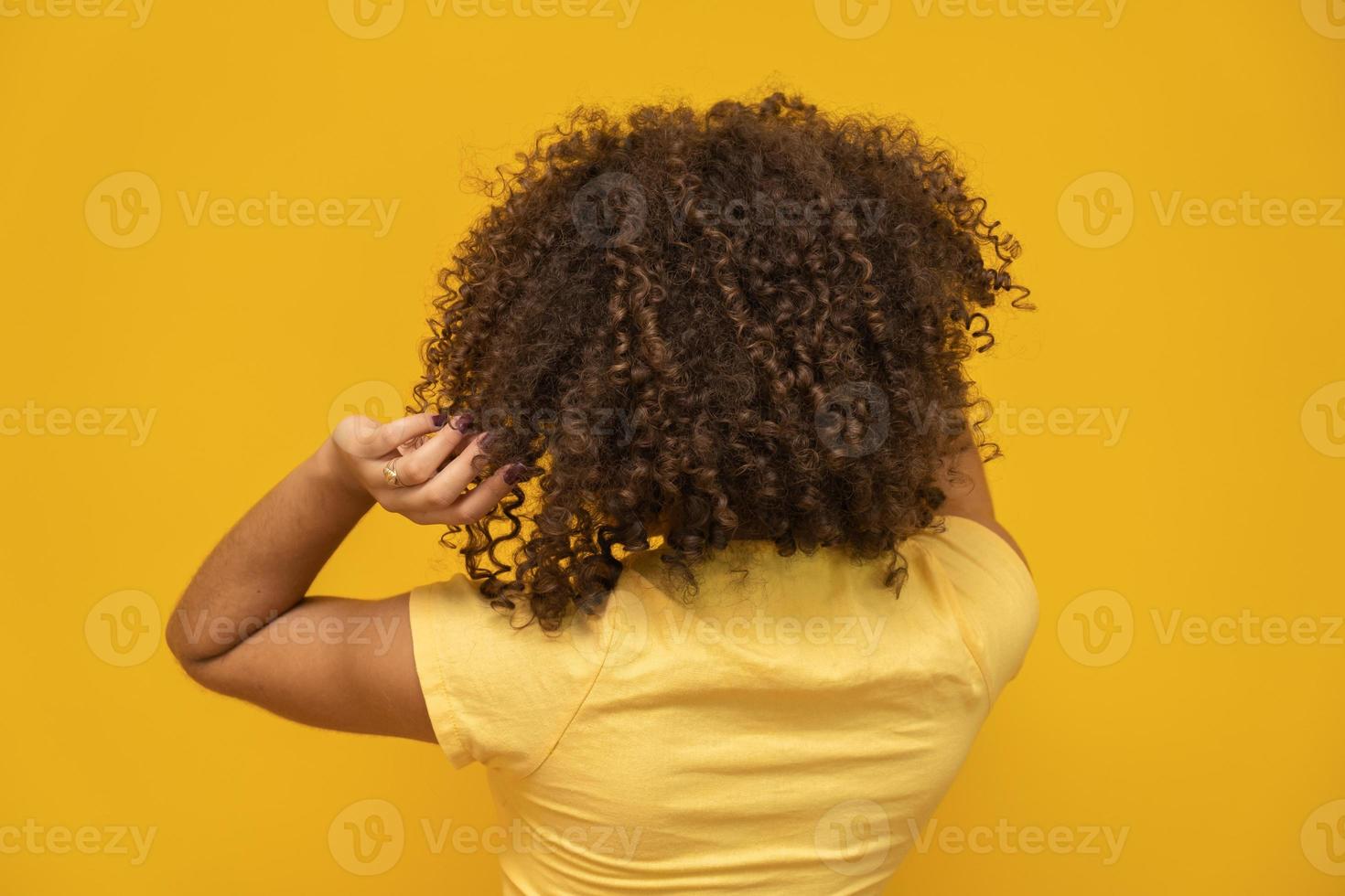 achteruit Amerikaanse Afrikaanse vrouw met haar krullend haar op gele achtergrond. lachende krullende vrouw in trui die haar haar aanraakt en naar de camera kijkt. foto