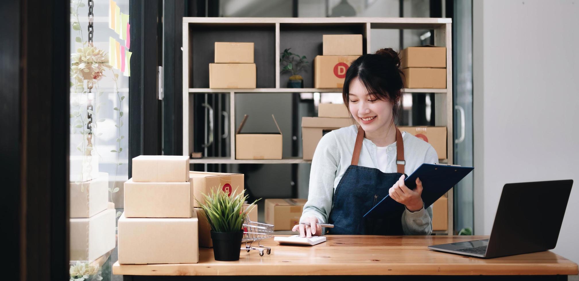 lachende jonge Aziatische bedrijfseigenaar vrouw bereidt pakketdoos voor en staande check online bestellingen van producten voor levering aan klant op laptopcomputer. online winkelconcept. foto