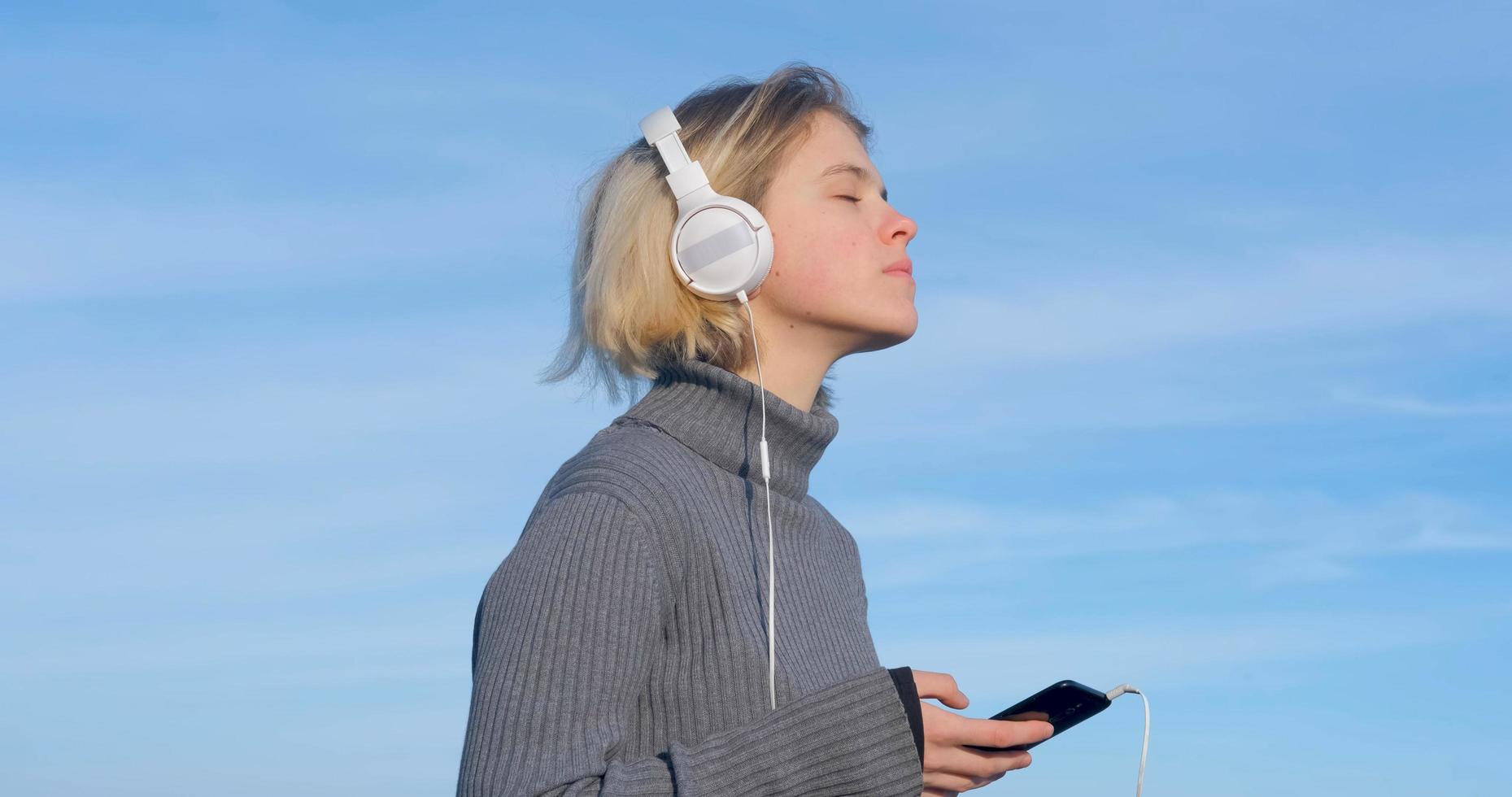 jonge knappe vrouw luistert naar muziek met een koptelefoon buiten op het strand tegen de zonnige blauwe hemel foto